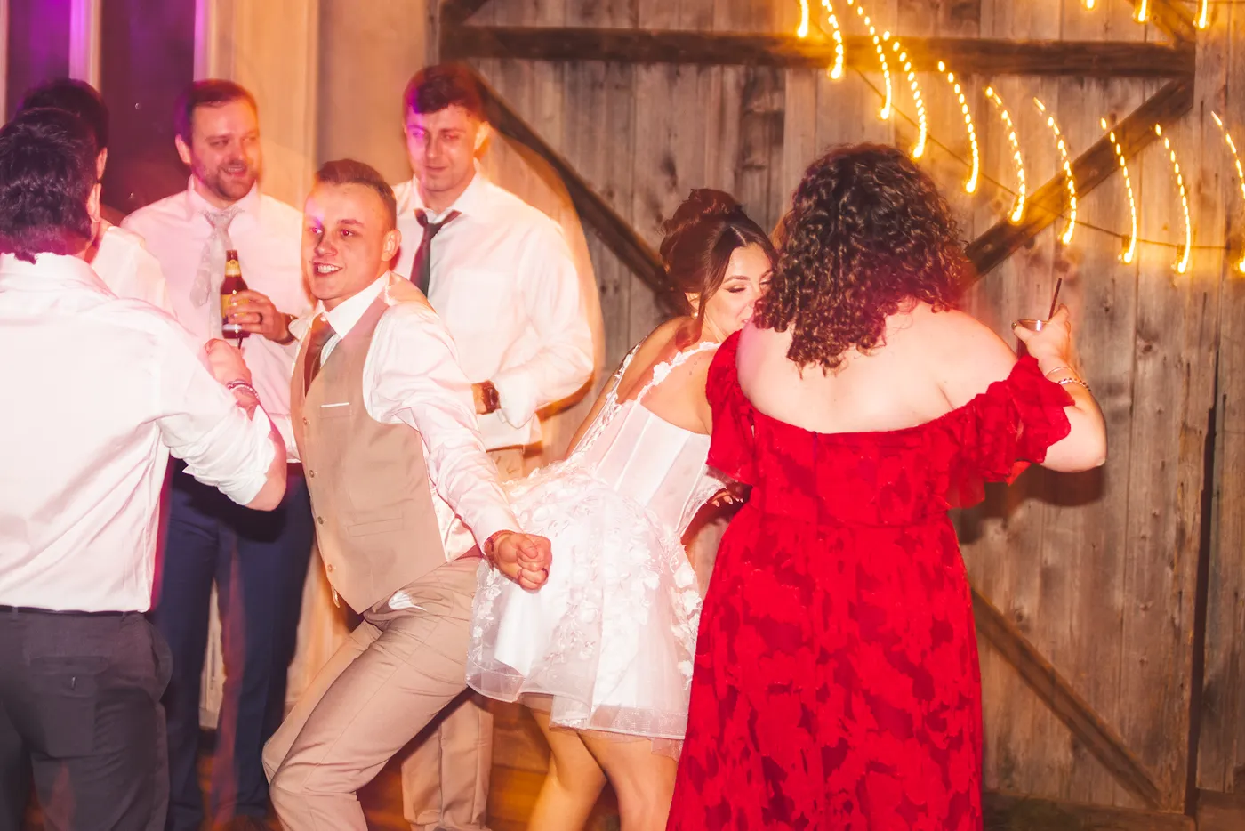 Guests dancing in a barn under warm string lights, the bride in the middle of them