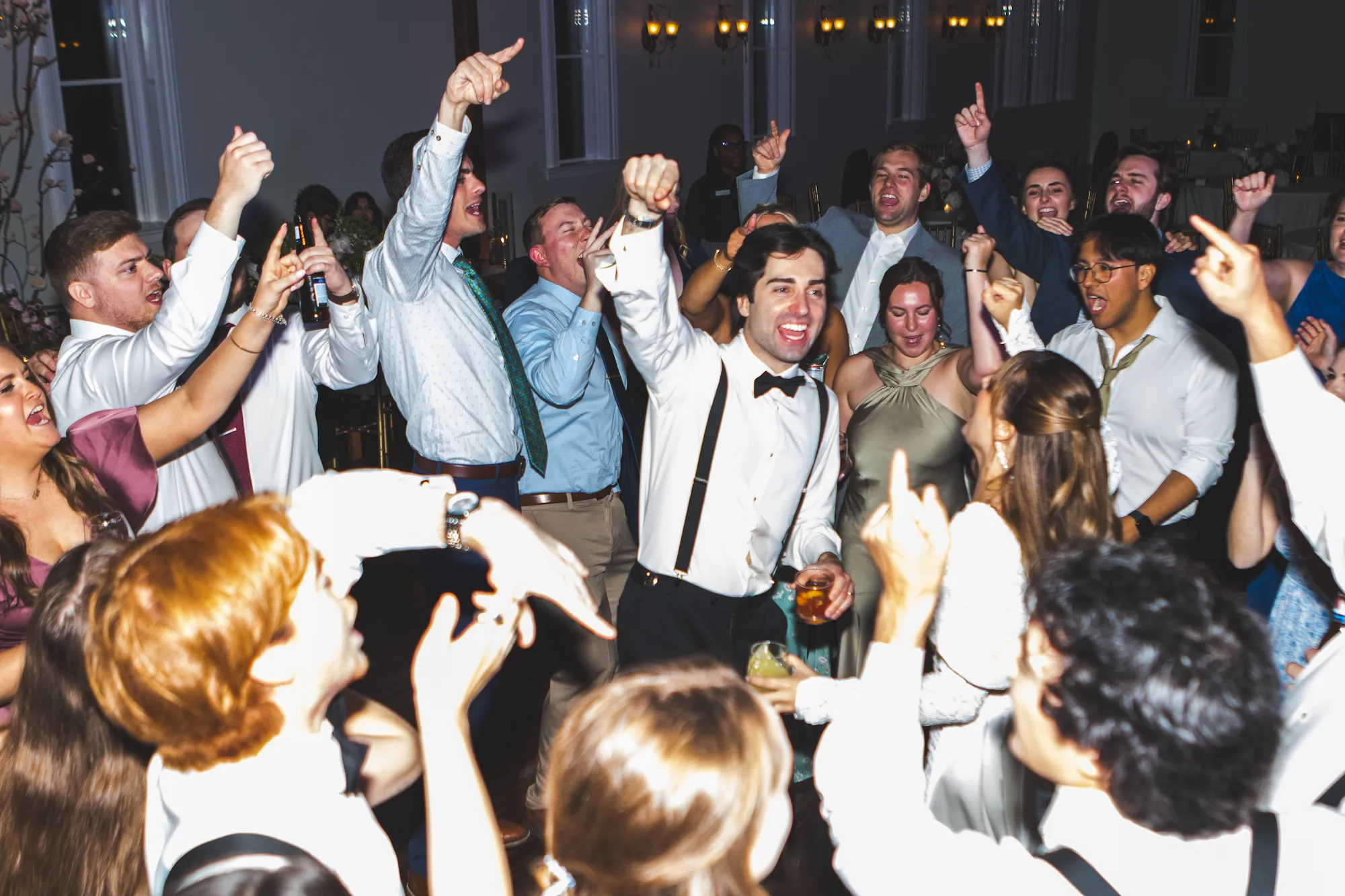 A packed wedding dance floor, the whole room cheering with their arms up under warm light