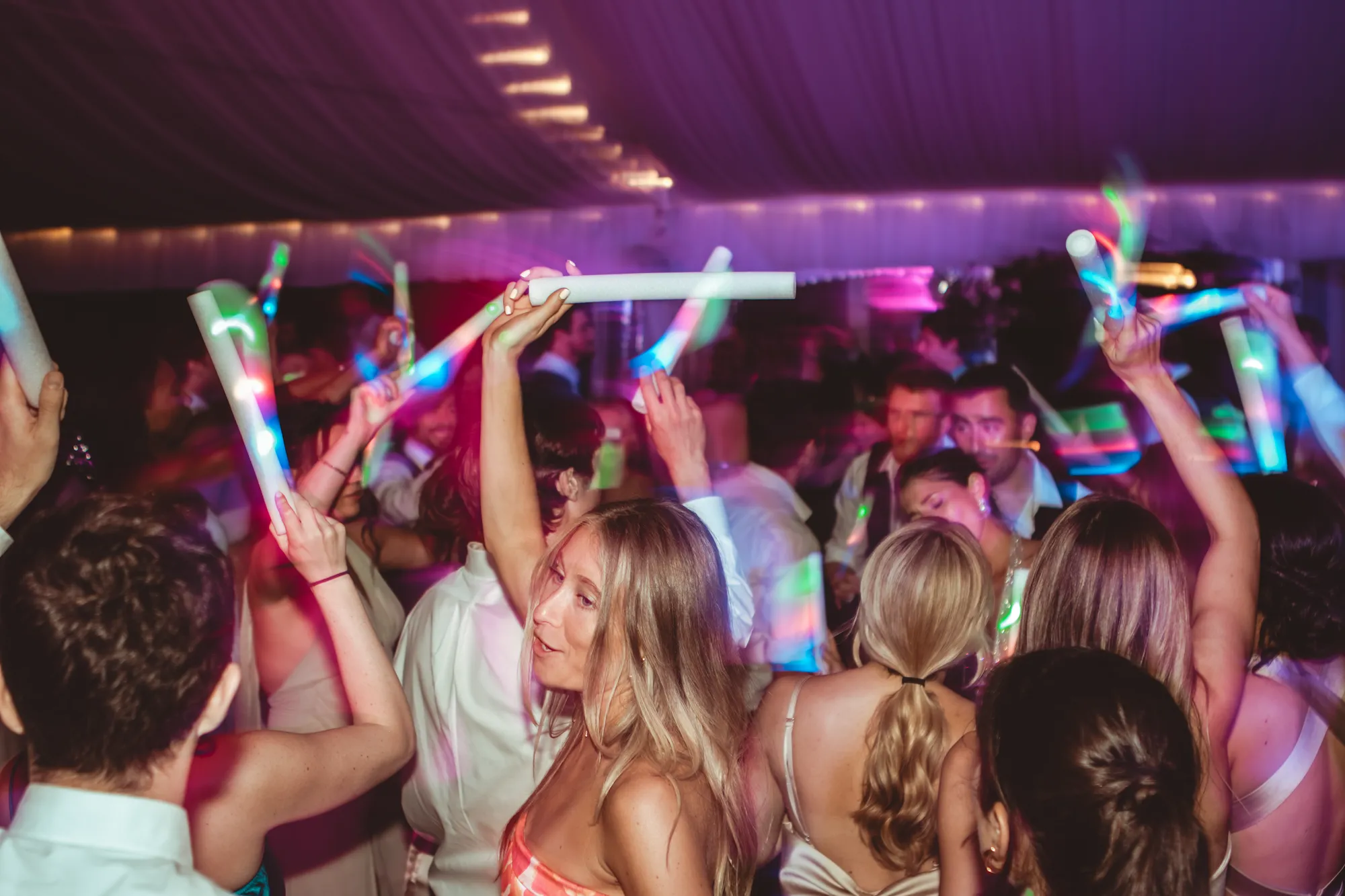 A packed tent dance floor washed in magenta, guests dancing with glow batons raised