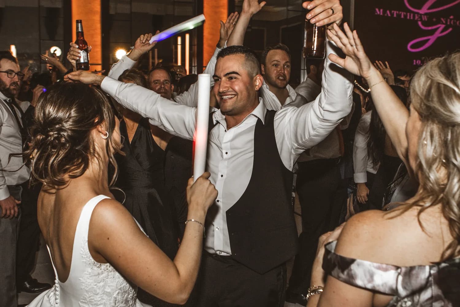A groom laughing mid-dance, surrounded by guests with their arms up, under warm light and a pink monogram Kyle projected on the back wall