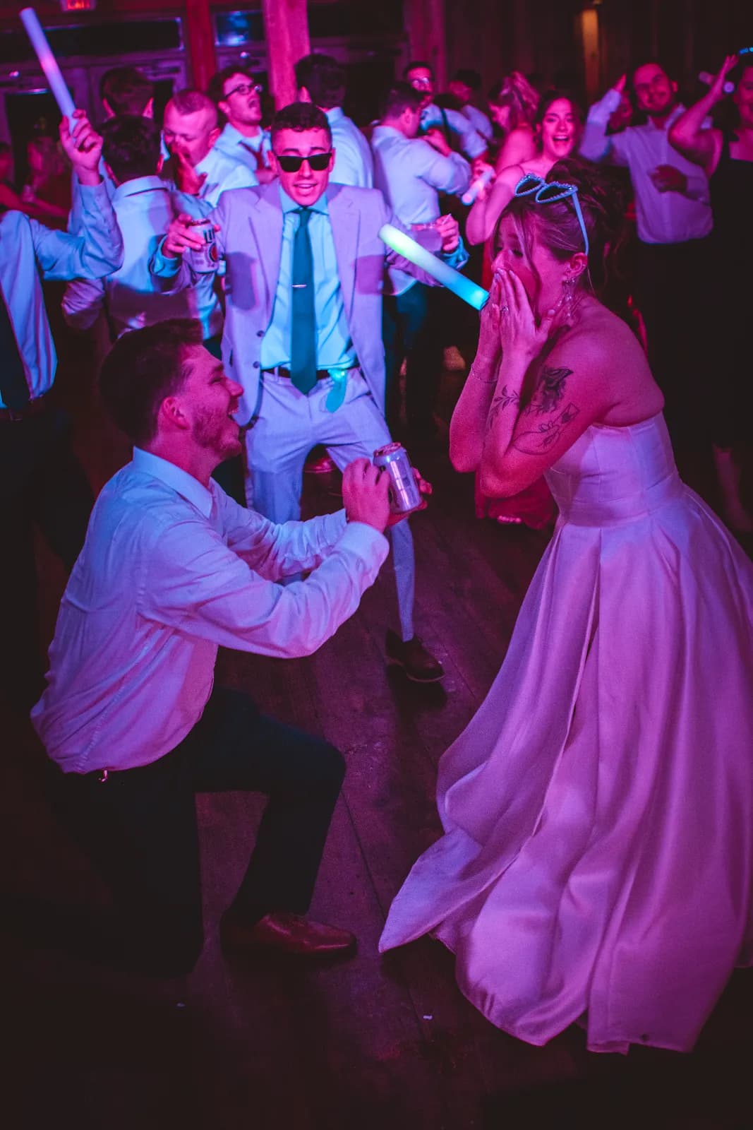 A bride laughing on a late-night dance floor lit in magenta and blue, a guest down on one knee in front of her